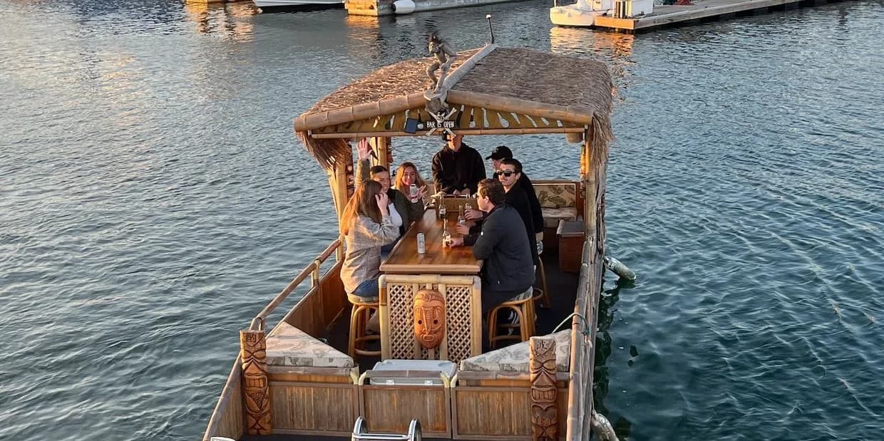 Tiki-themed party boat with canopy and bar, passengers seated on deck over water