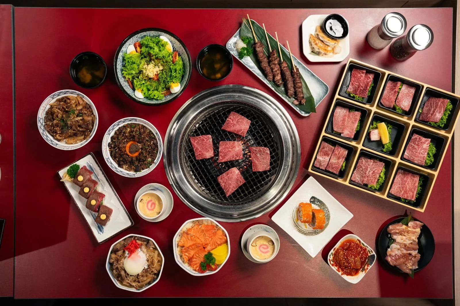 Wagyu beef dishes arranged overhead: tabletop grill with sliced meat, grilled items, salads, and side dishes on red surface