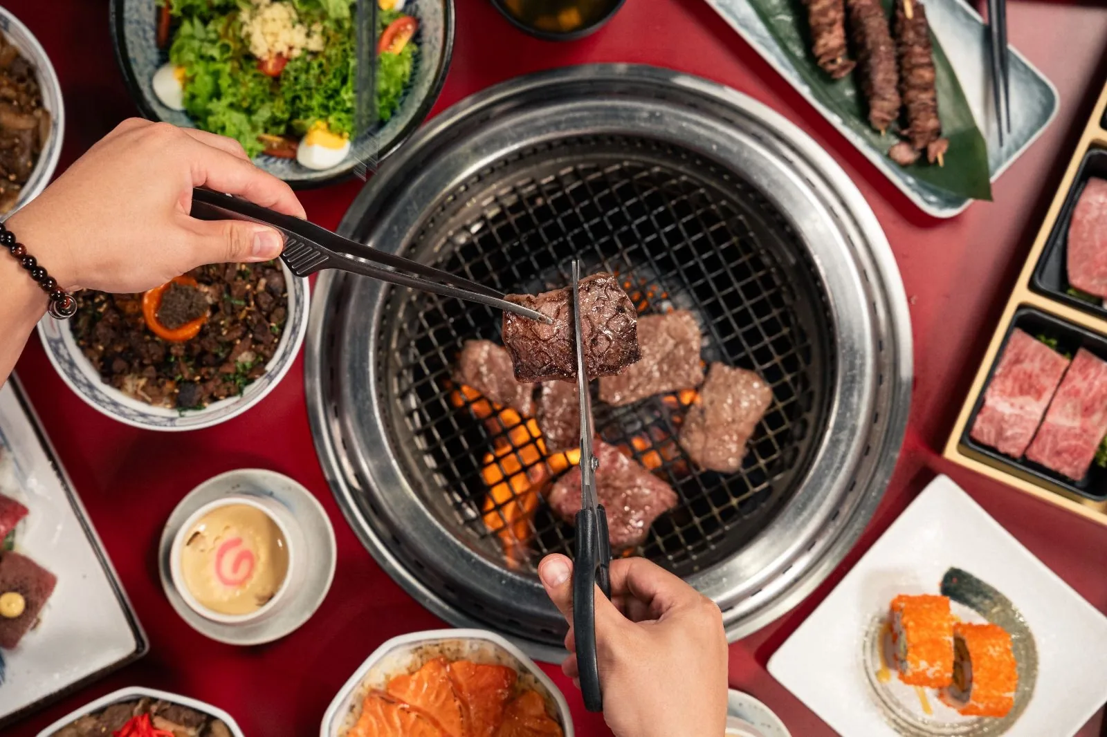 Hands grilling meat on a tabletop Korean BBQ grill with side dishes and ingredients around the table