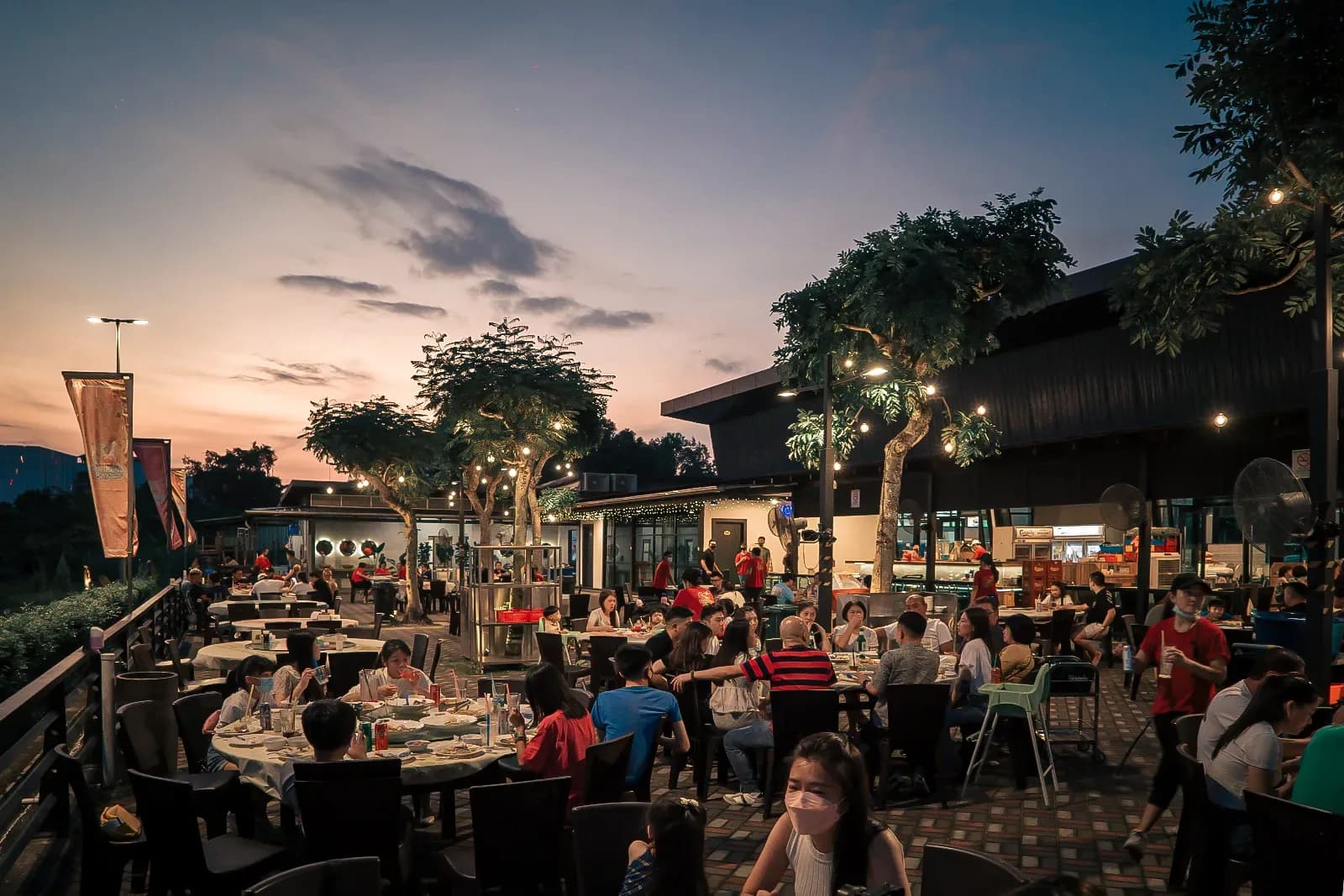 Outdoor seafood restaurant patio at dusk with diners at tables under trees and mood lighting.