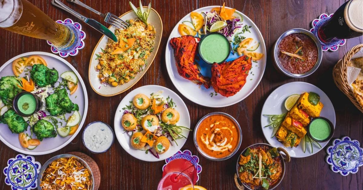 Array of North Indian curries, tandoori meats, breads, and dips arranged on white plates with blue ceramic coasters on dark wood table