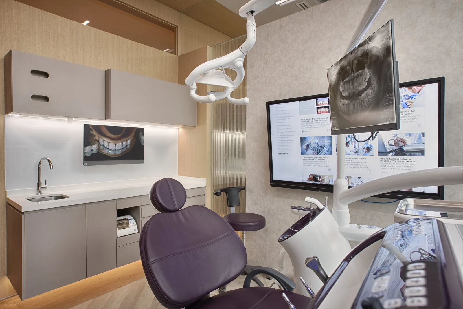 Modern dental treatment room with purple chair, overhead light, and monitors displaying X-rays