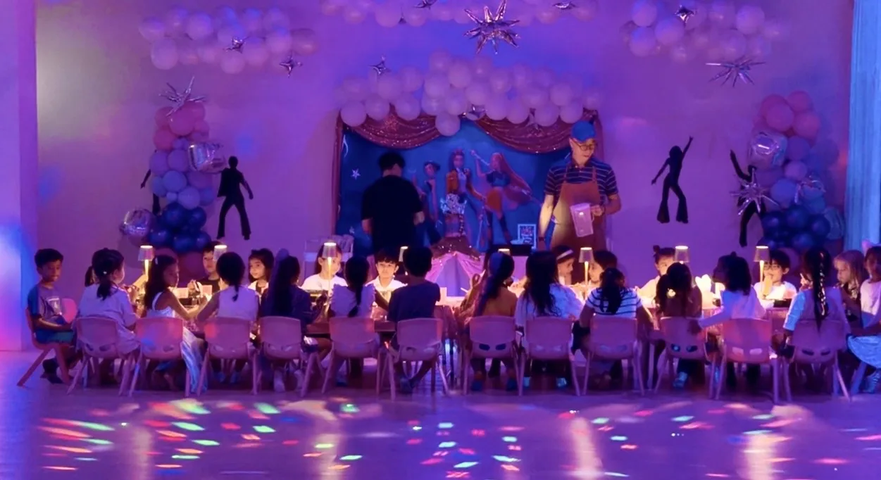 Kids' birthday party with children at tables, lit stage with performers, colorful purple lighting and decorations.