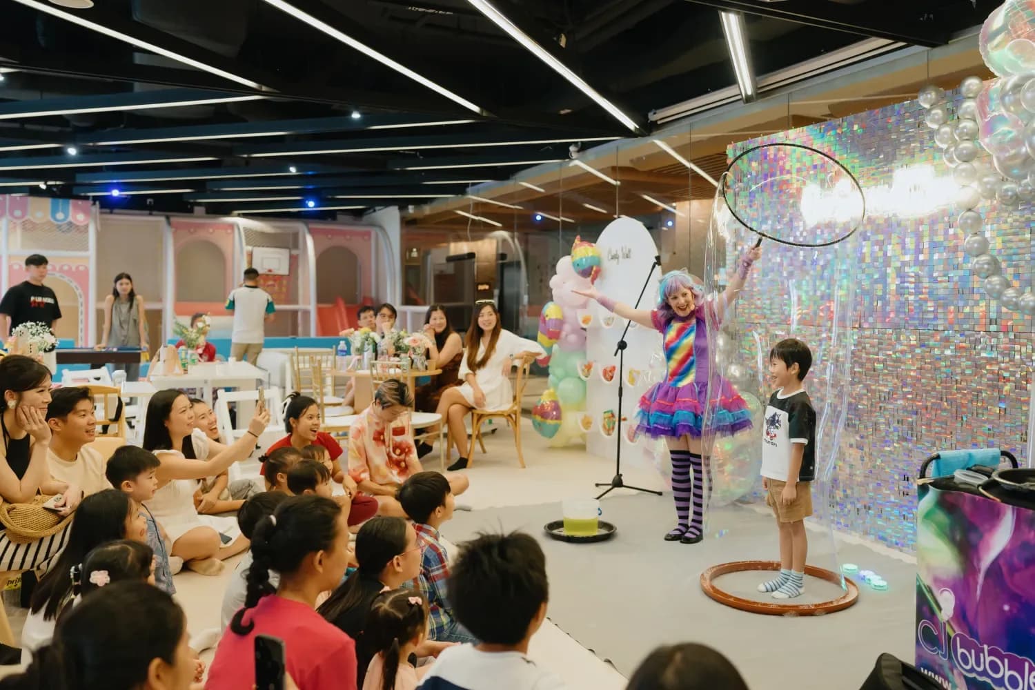 Children watch an entertainer in a rainbow costume perform at a birthday party in a modern event space.