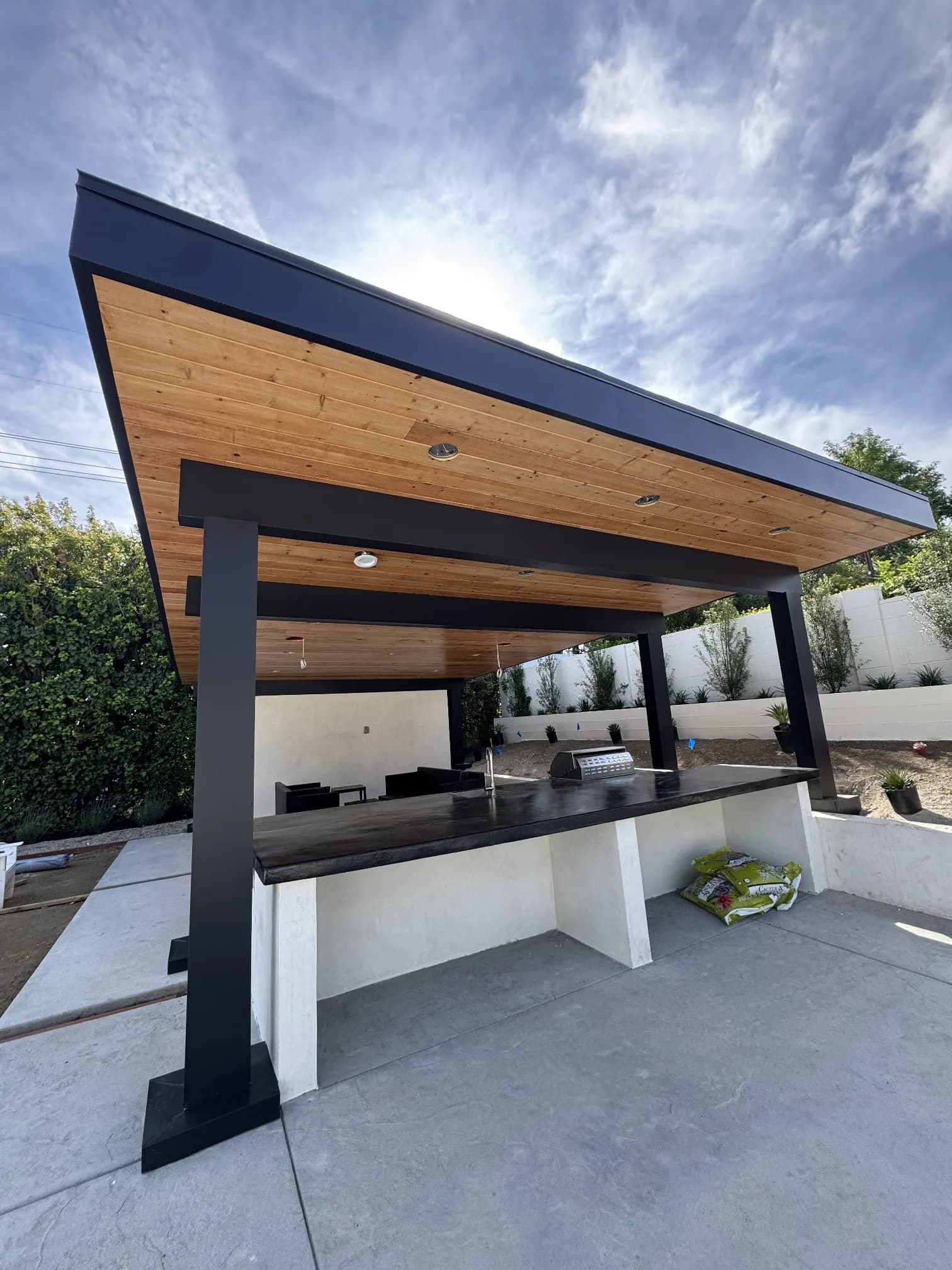 Modern metal-framed outdoor kitchen shelter with wood ceiling and built-in counter