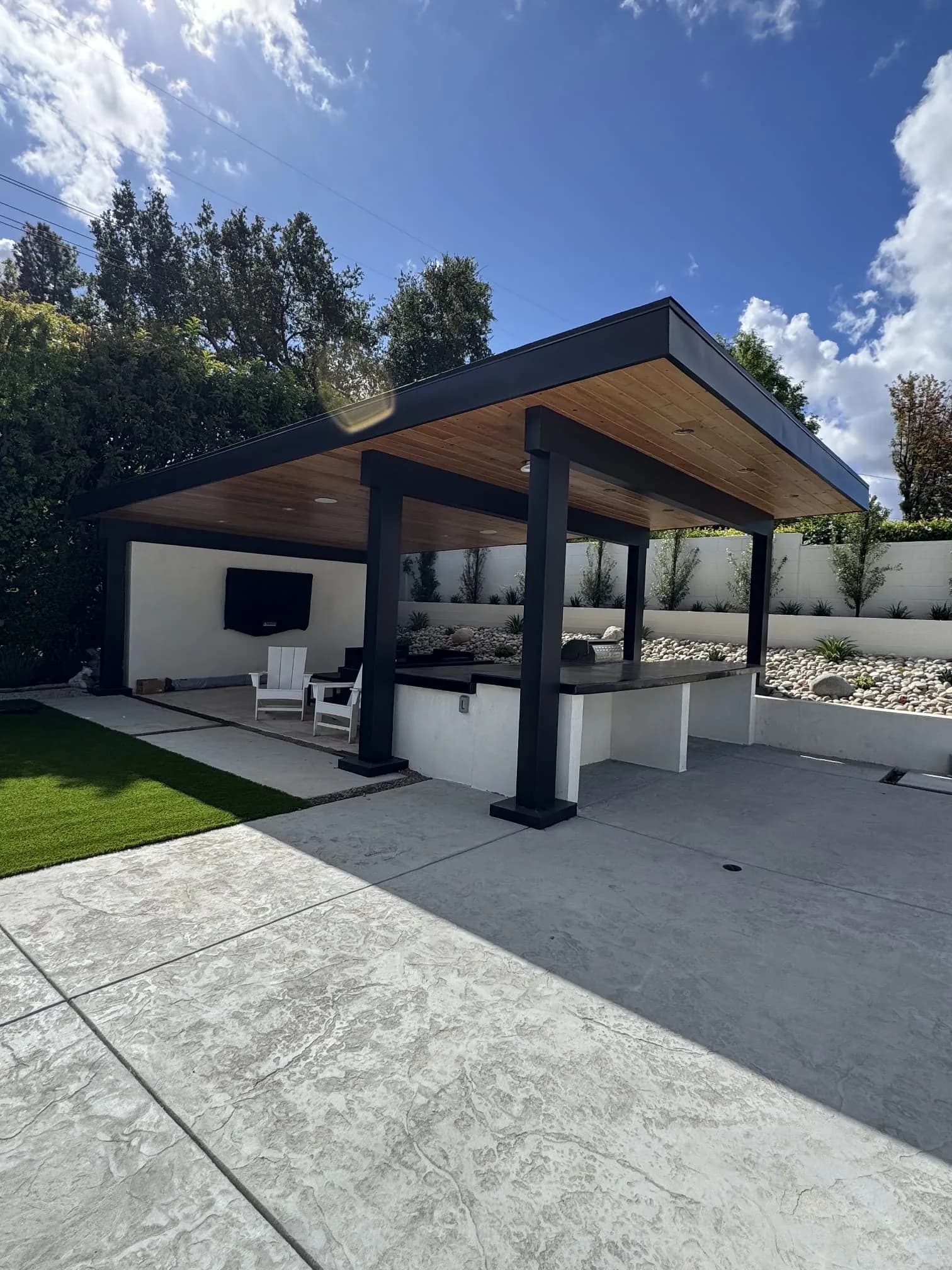 Modern patio with black metal frame, wood soffit, white built-ins, and concrete flooring in landscaped yard