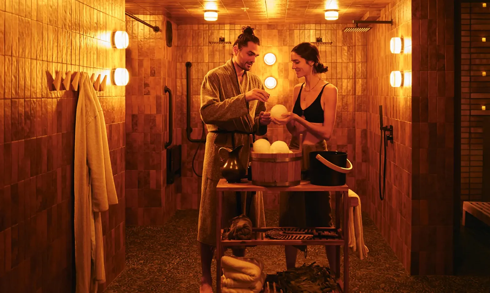 Two people in a wooden sauna with glowing lights and heated stones on a table