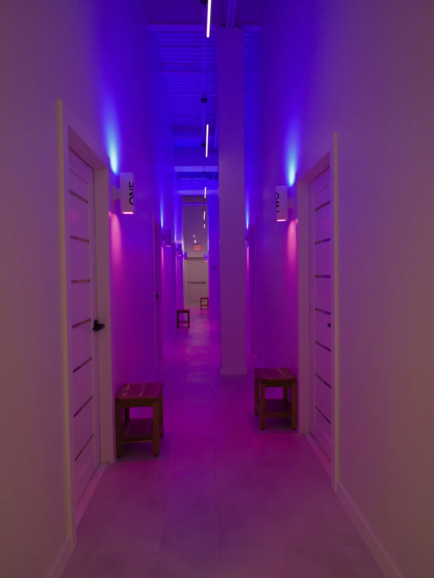 Neon-lit hallway in a bathhouse with purple and blue lighting, white doors, and modern aesthetic