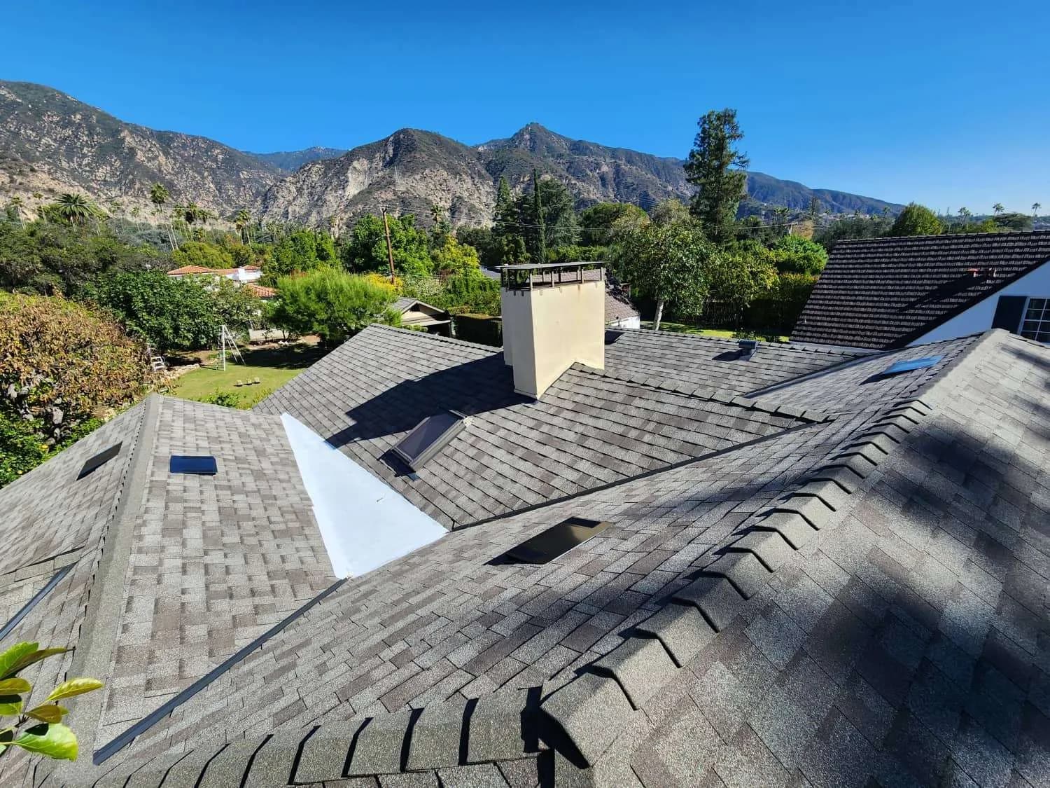 Newly installed dark asphalt shingle roof with white skylight opening, chimney, and mountain landscape in background.