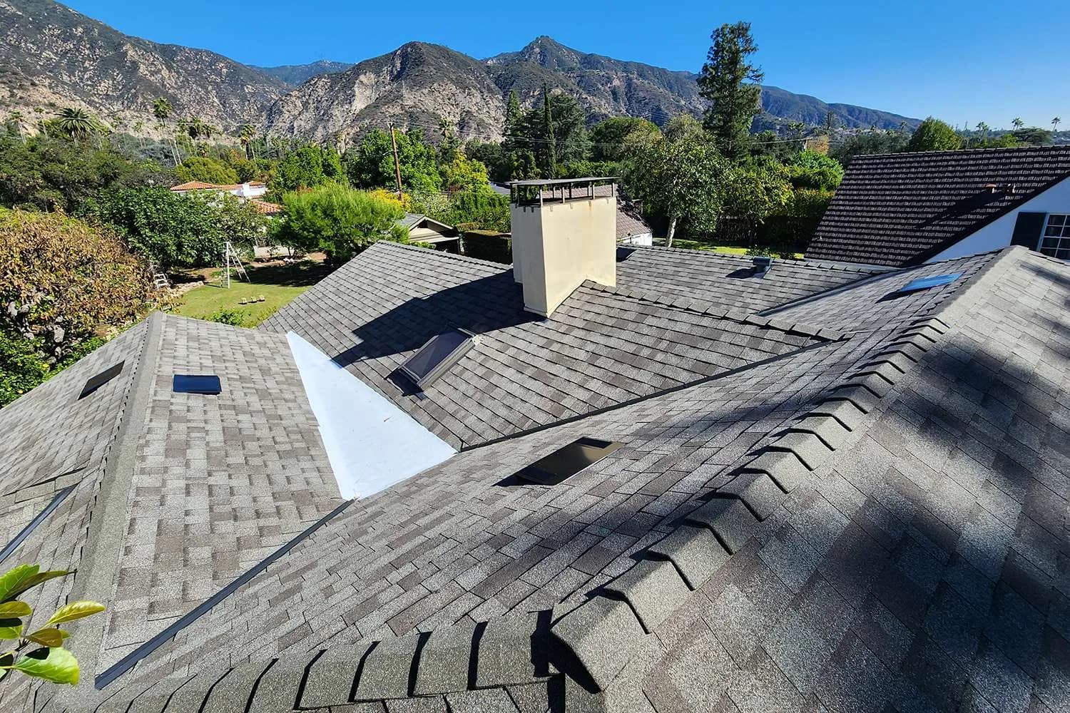 Residential roof installation with new gray shingles, white flashing, and chimney in foothills setting
