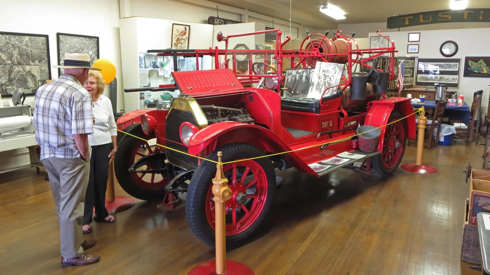 Vintage red fire truck on display in a museum with two visitors viewing it, surrounded by historical photos and memorabilia