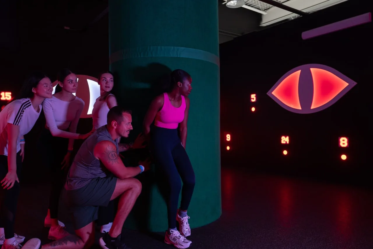 Group of people participating in an active game or workout in a neon-lit entertainment venue with scoring display.