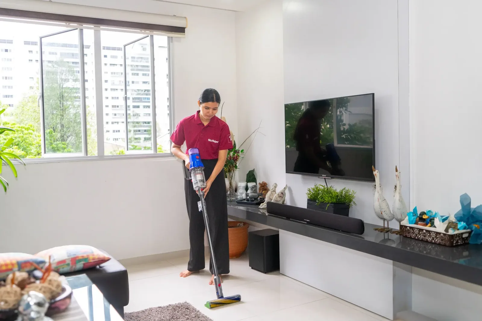 Cleaner vacuuming a bright living room with city view windows in the background