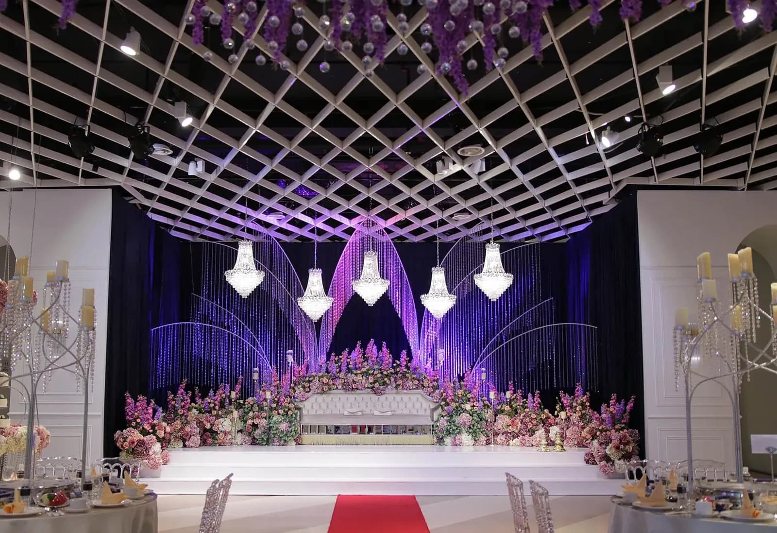 Grand wedding venue stage with crystal chandeliers, purple lighting, and pink floral arrangements