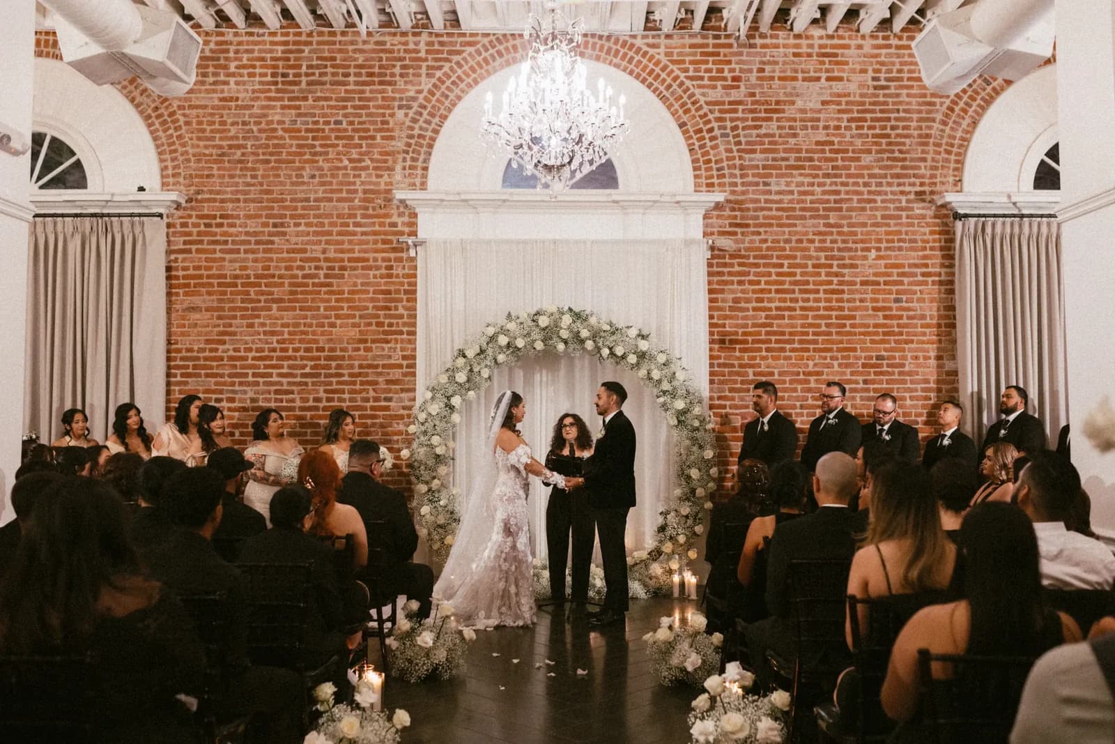 Wedding ceremony in brick venue with floral arch, guests seated, and crystal chandelier