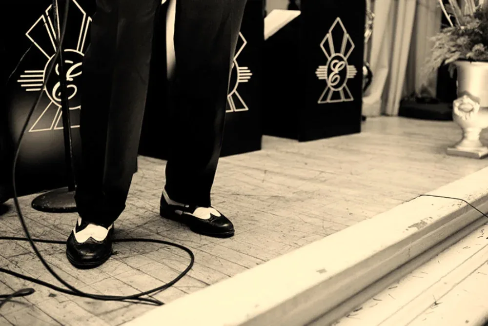 Performers in formal black outfits with geometric embroidery stand on wooden flooring during an event.
