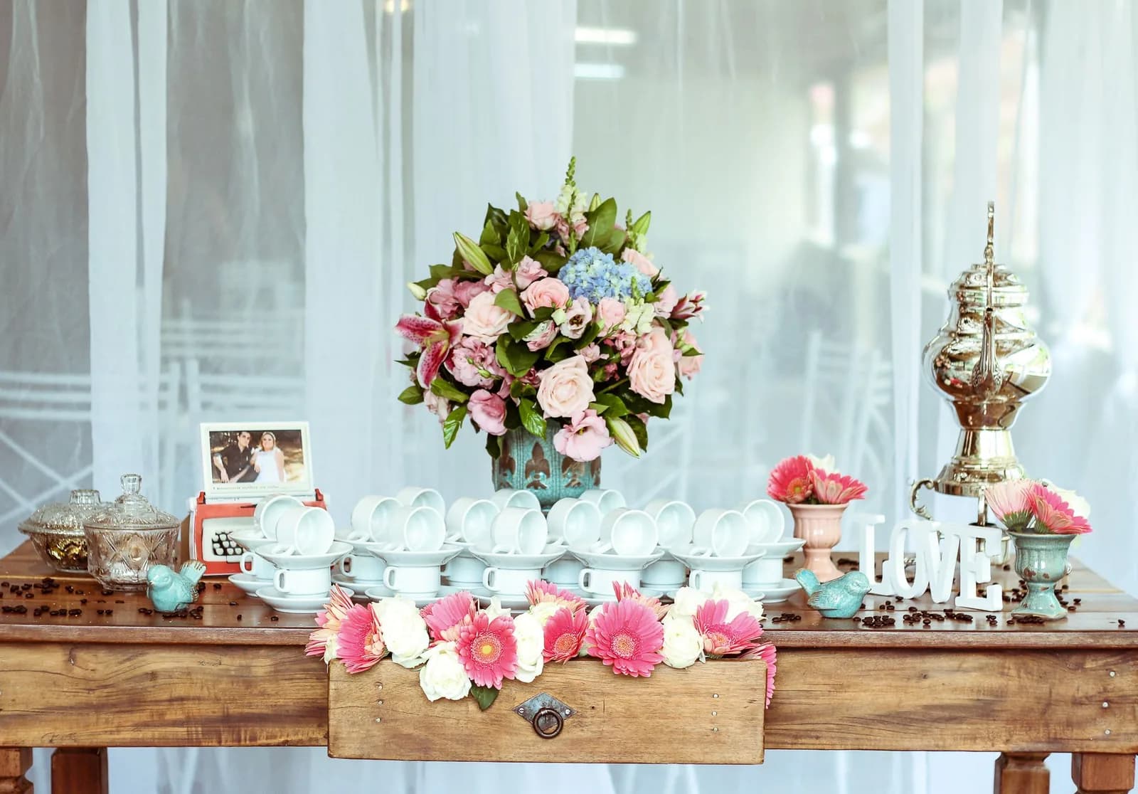 Styled event display with pink and blue floral arrangements, white china, and vintage decorative pieces on wooden table.