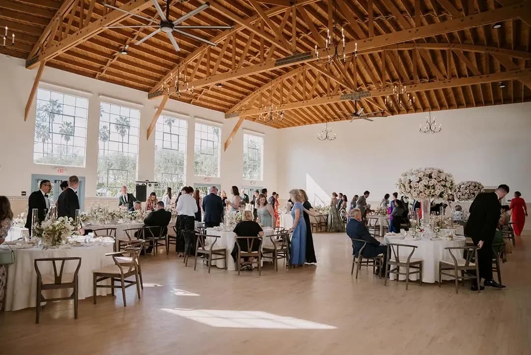 Elegant event venue with wooden beams, white walls, round tables set for dining, and guests at a reception