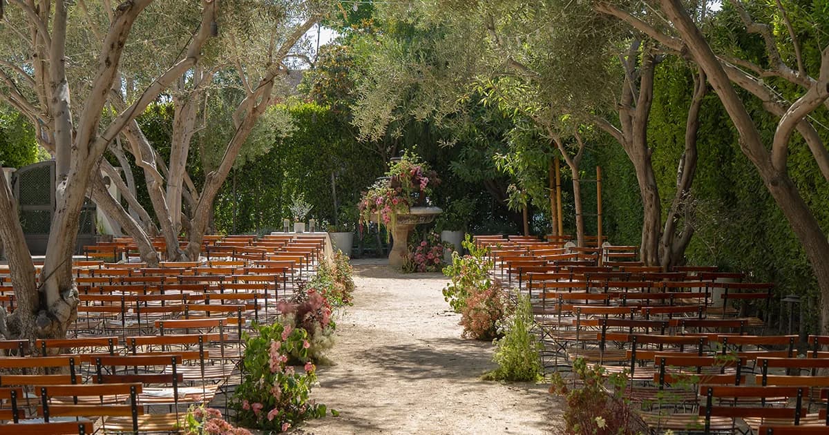 Wooden chairs arranged under tall eucalyptus trees with pink floral decorations and a stone urn