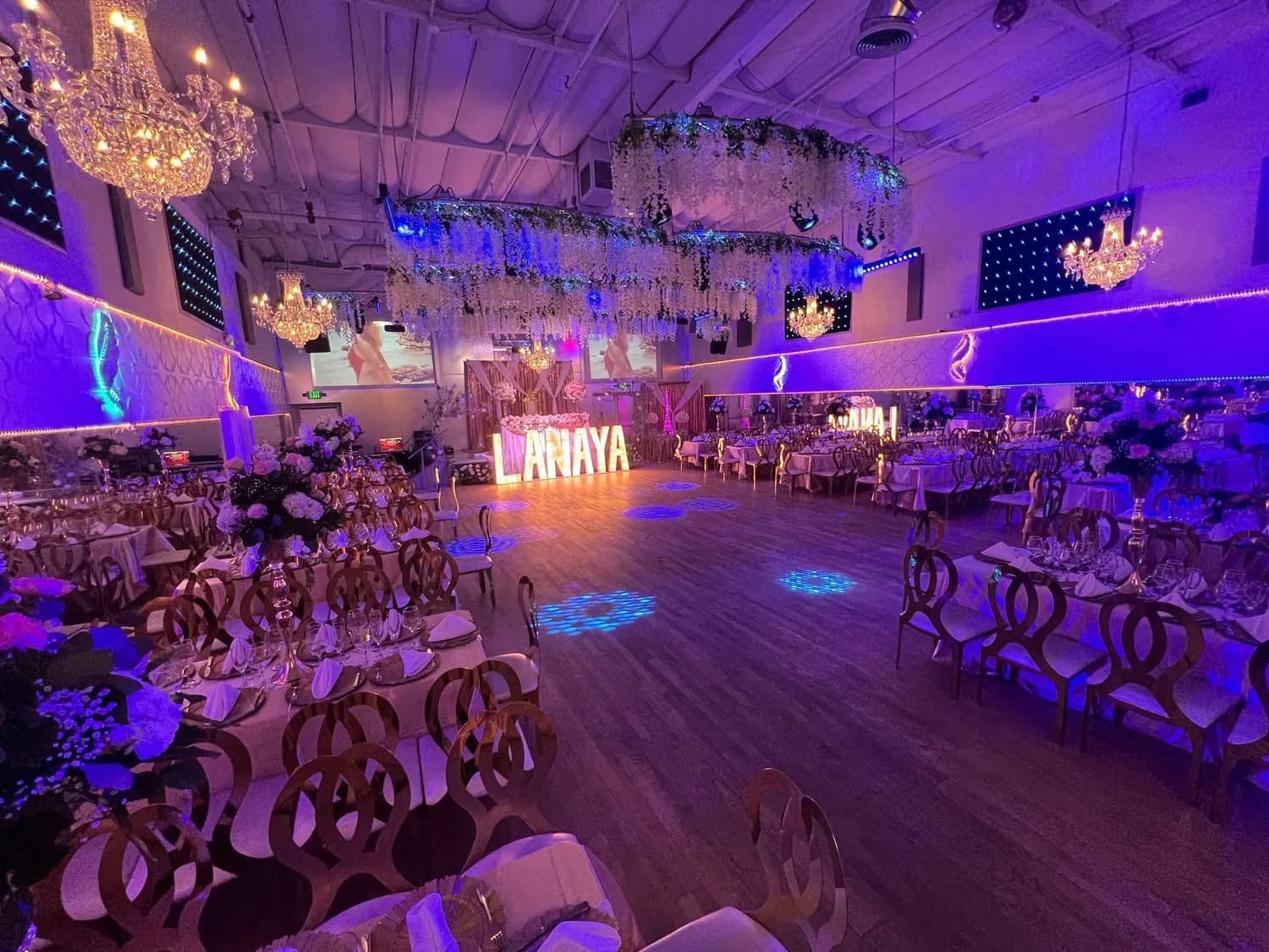 Upscale event venue with purple lighting, crystal chandeliers, round tables, and illuminated dance floor