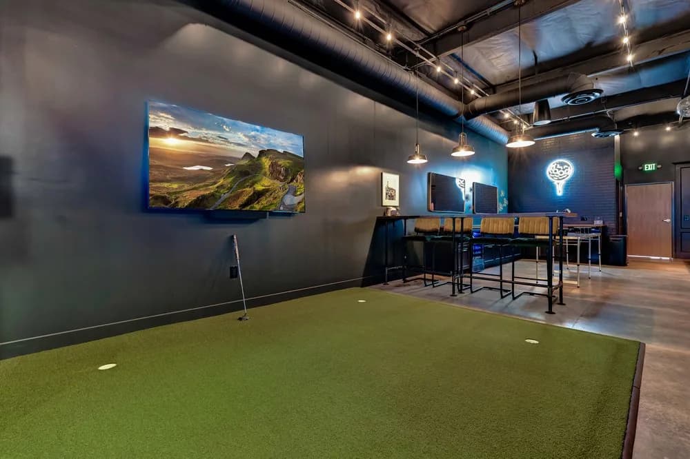 Indoor golf simulator and bar area with green putting surface, high-top seating, and neon signage