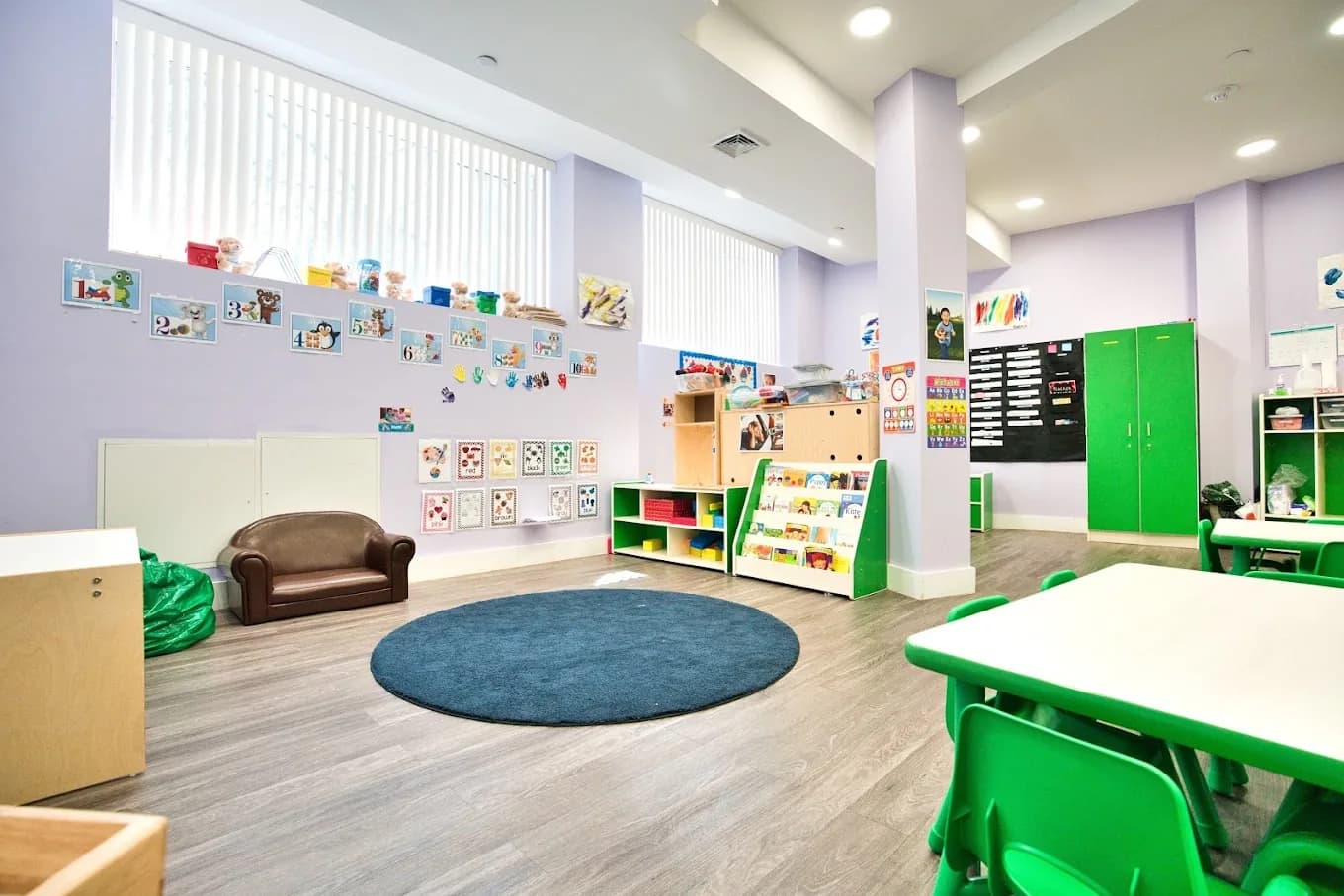 Bright preschool classroom with green furniture, blue rug, and educational displays on purple walls.