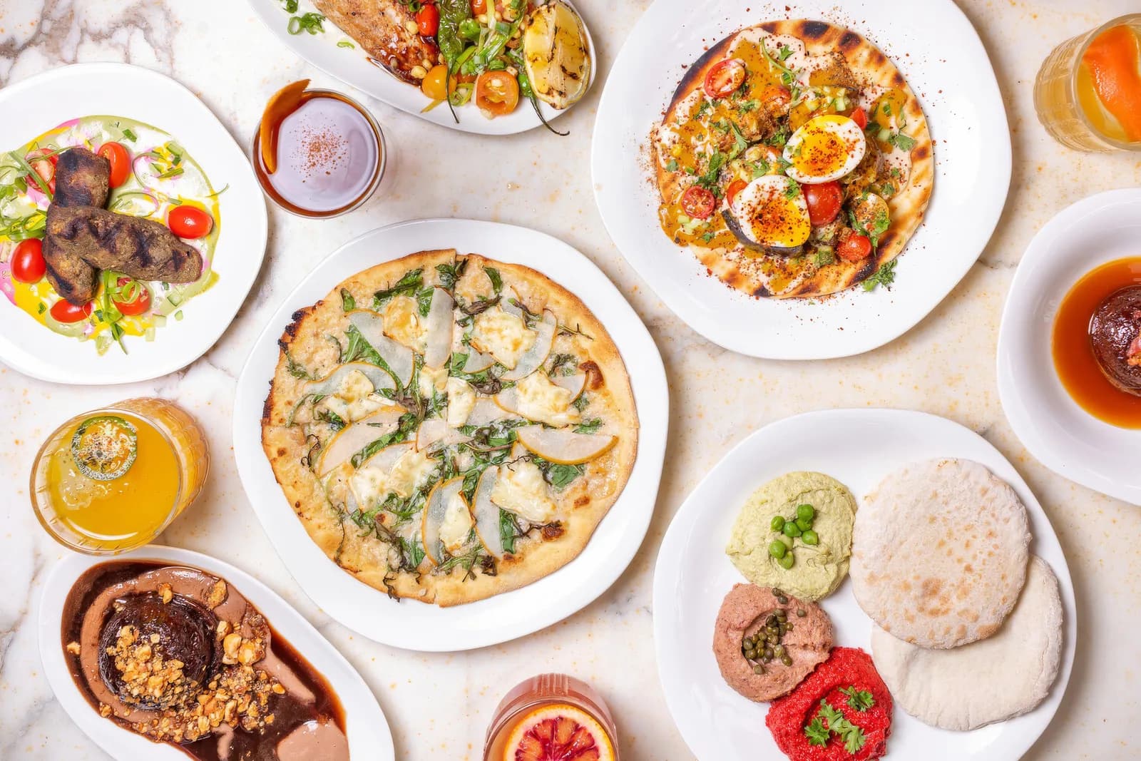 Overhead view of restaurant dishes: pizzas, grilled meat, appetizers, and desserts with drinks on marble surface.