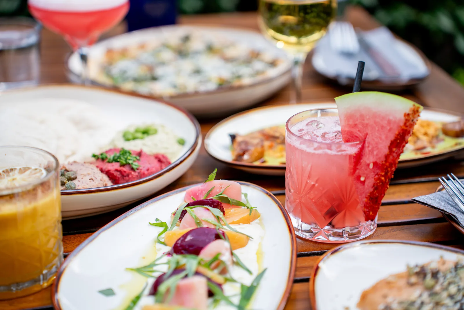 Outdoor restaurant table with colorful plated dishes, pink cocktail, and wine glasses