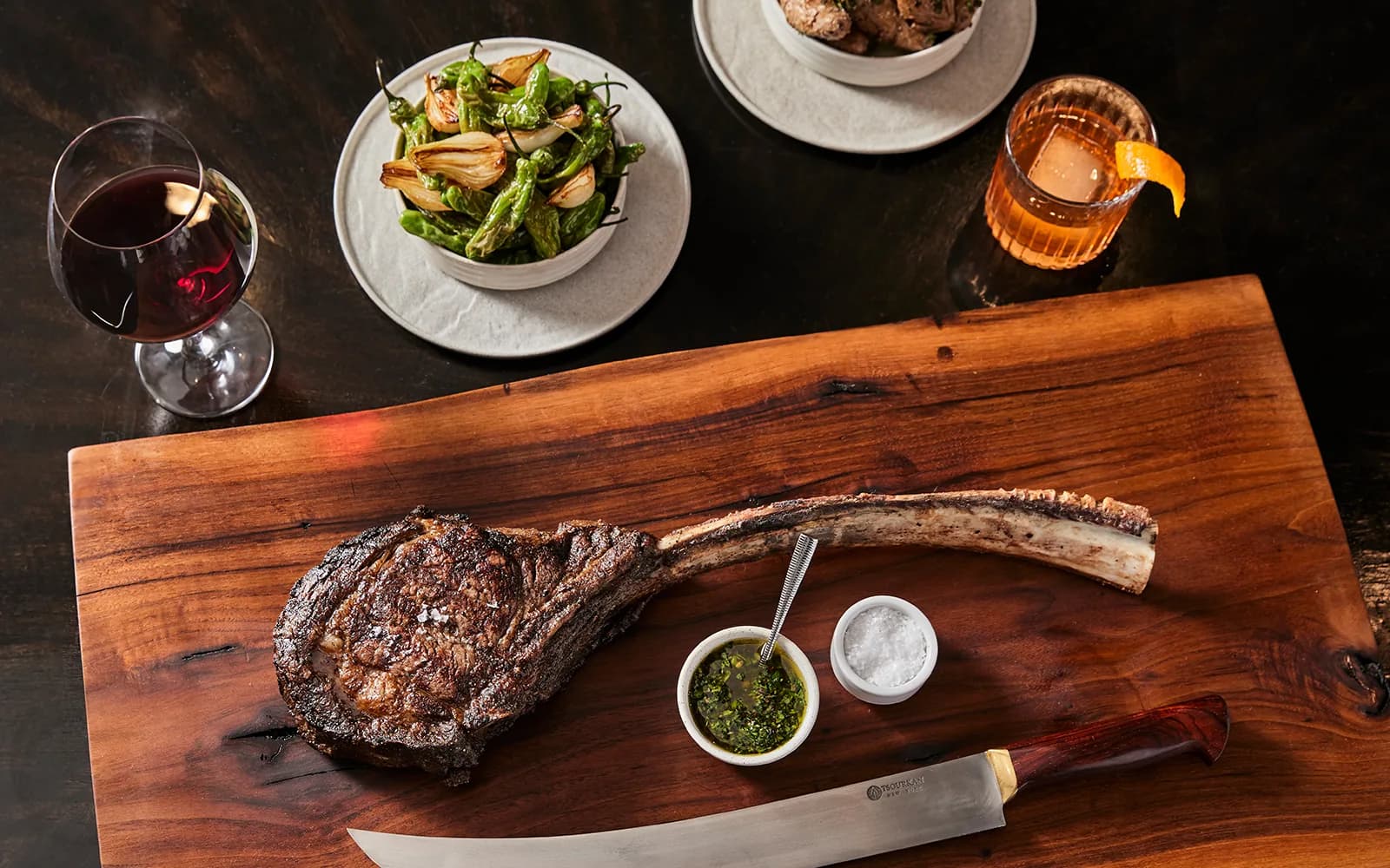 Grilled tomahawk steak on wooden board with chimichurri, salt, and knife, plus wine and appetizers.