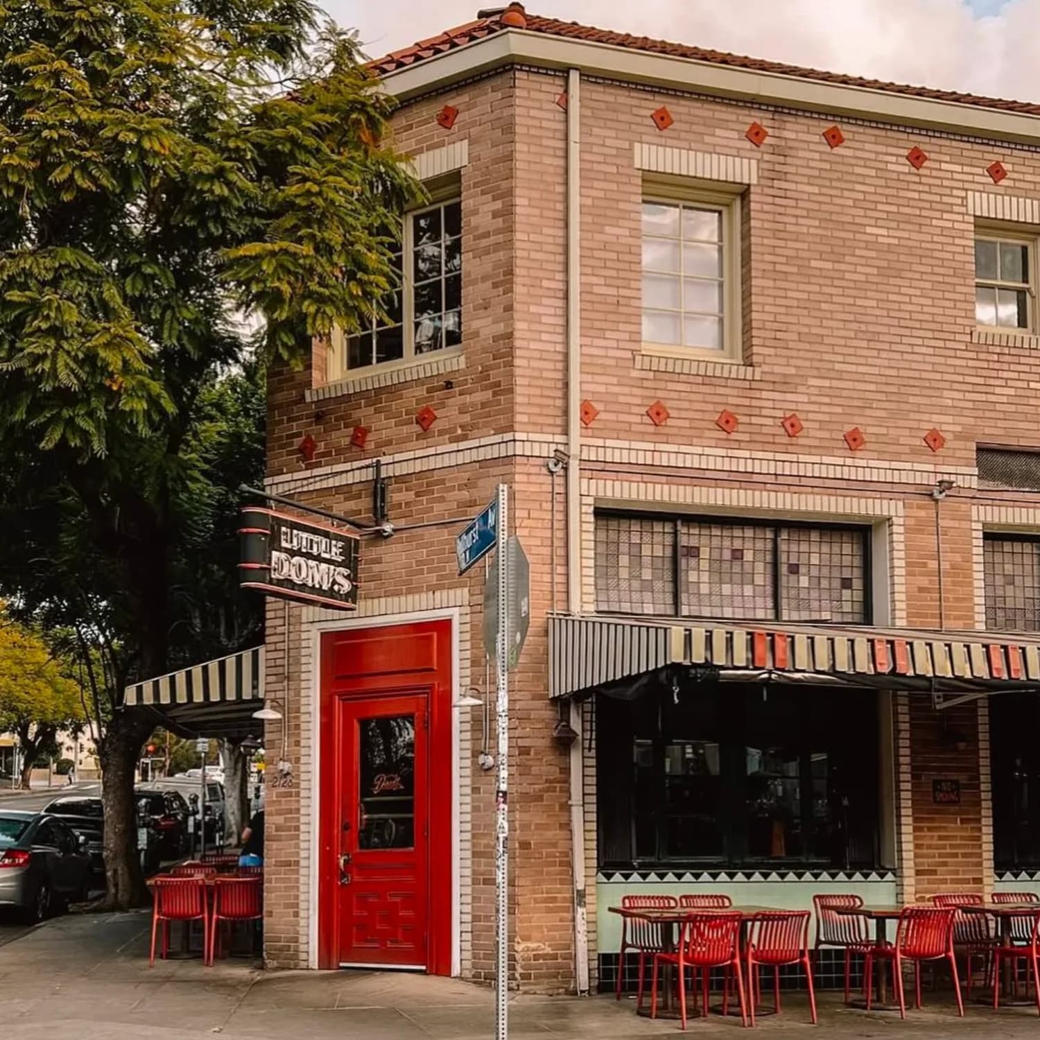 Red brick building exterior with red door, storefront windows, outdoor seating with red chairs, and signage