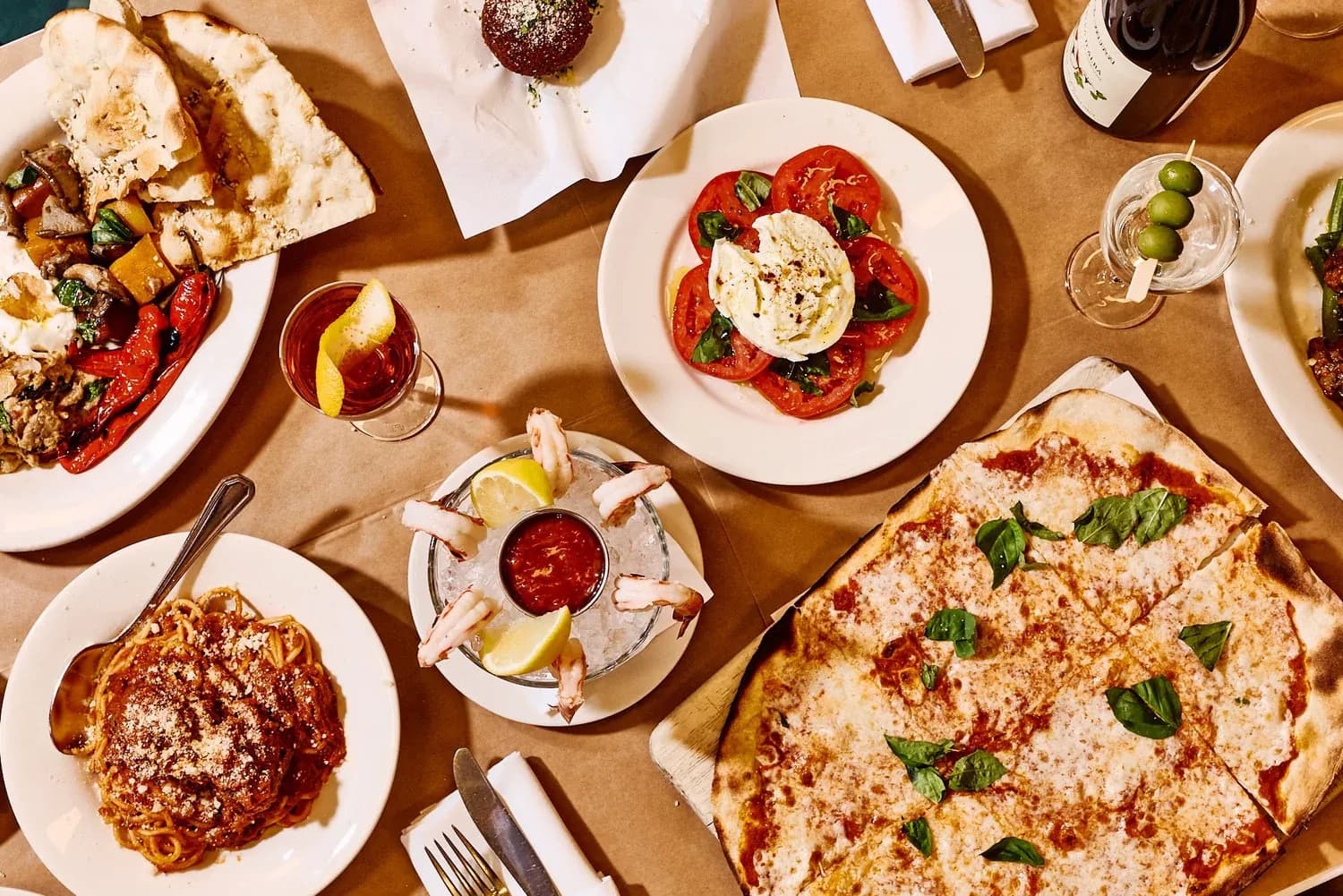 Overhead view of Italian restaurant dishes including pizza, caprese, pasta, and cocktails on a wood table