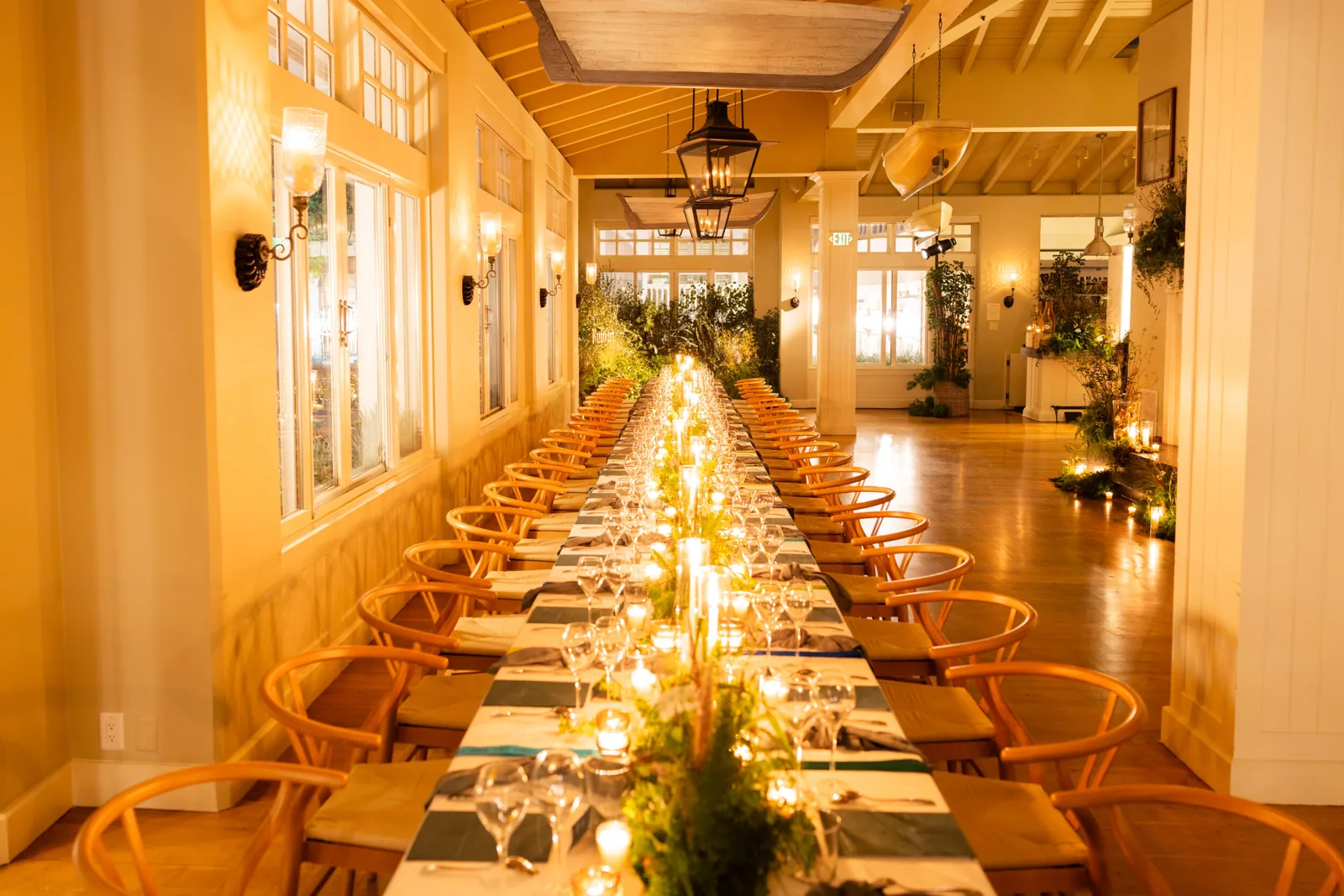 Long dining table set for an event with wooden chairs, place settings, candles, and greenery decorations