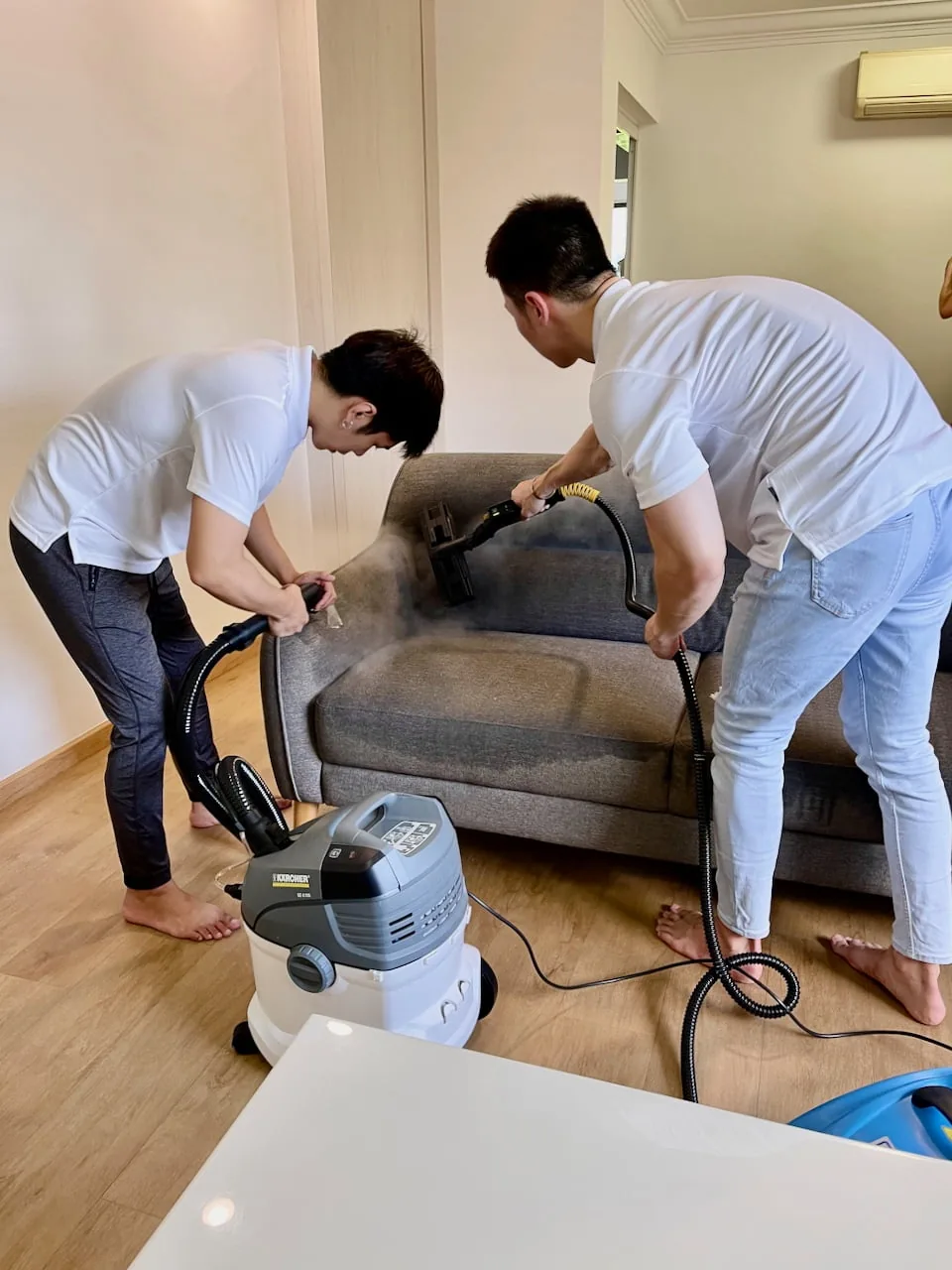 Two workers cleaning a grey sofa with professional steam cleaning machine in a home interior.