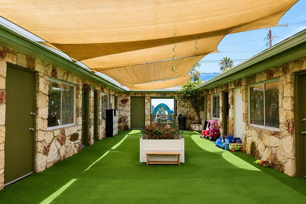 Preschool outdoor classroom with stone walls, shade sails, green turf, and toys under clear sky