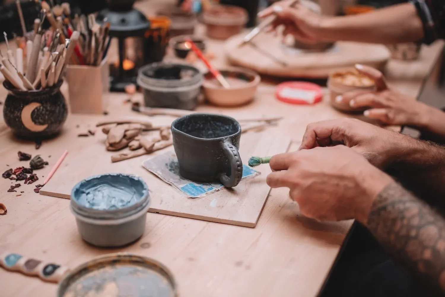 Pottery studio participants painting and decorating ceramic pieces at a wooden work table with supplies