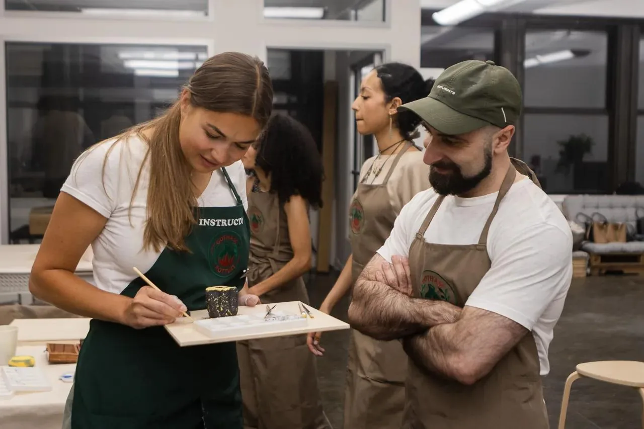 Pottery instructor demonstrates hand-building technique to student at studio workstation