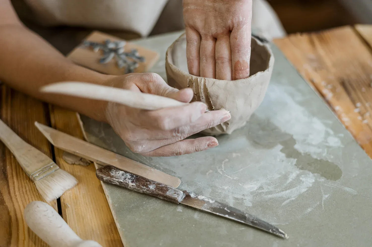 Hands shaping clay during a pottery demonstration, with brushes and tools on wooden table.