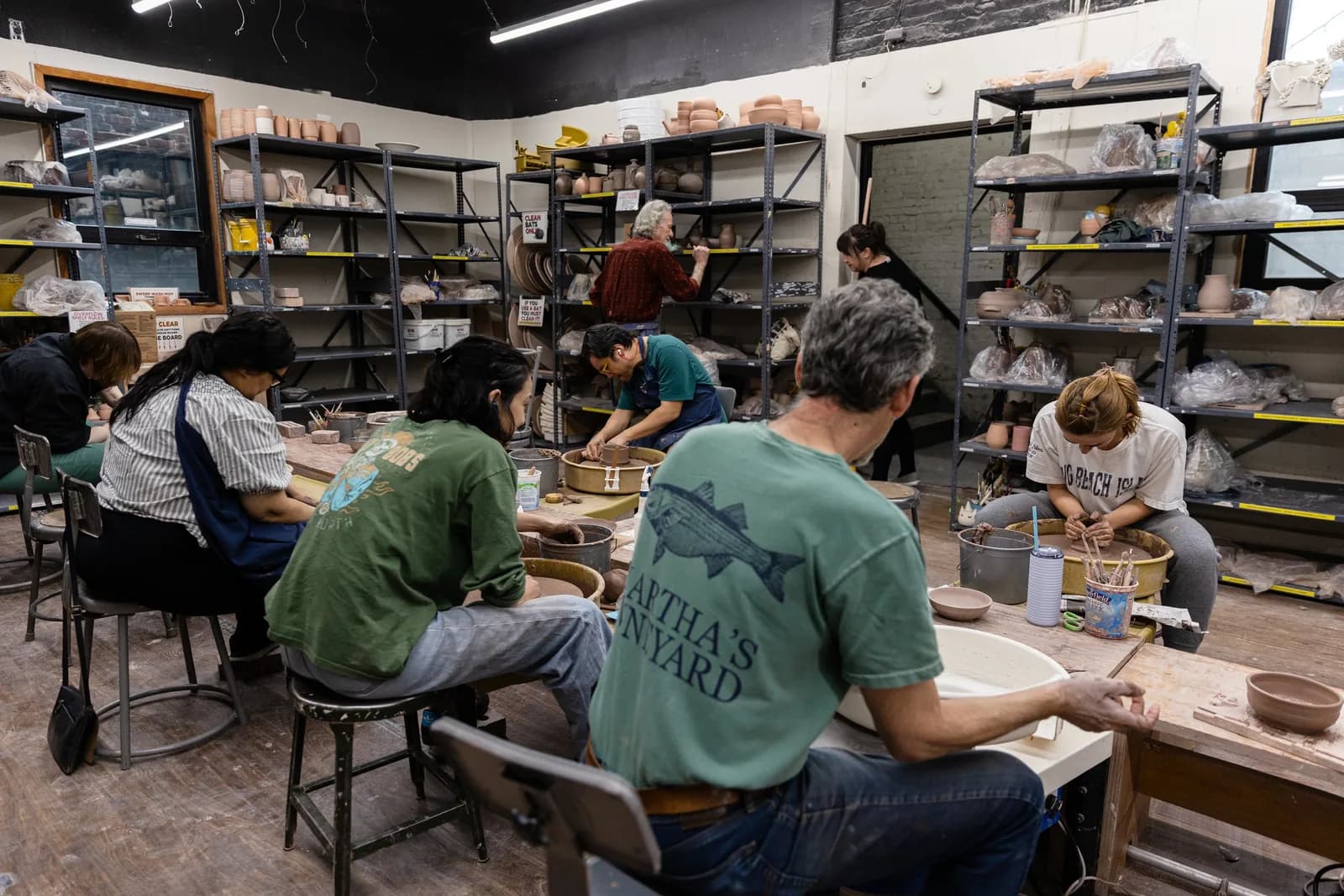 Pottery students working at tables in a ceramic studio surrounded by shelves of clay vessels and supplies.
