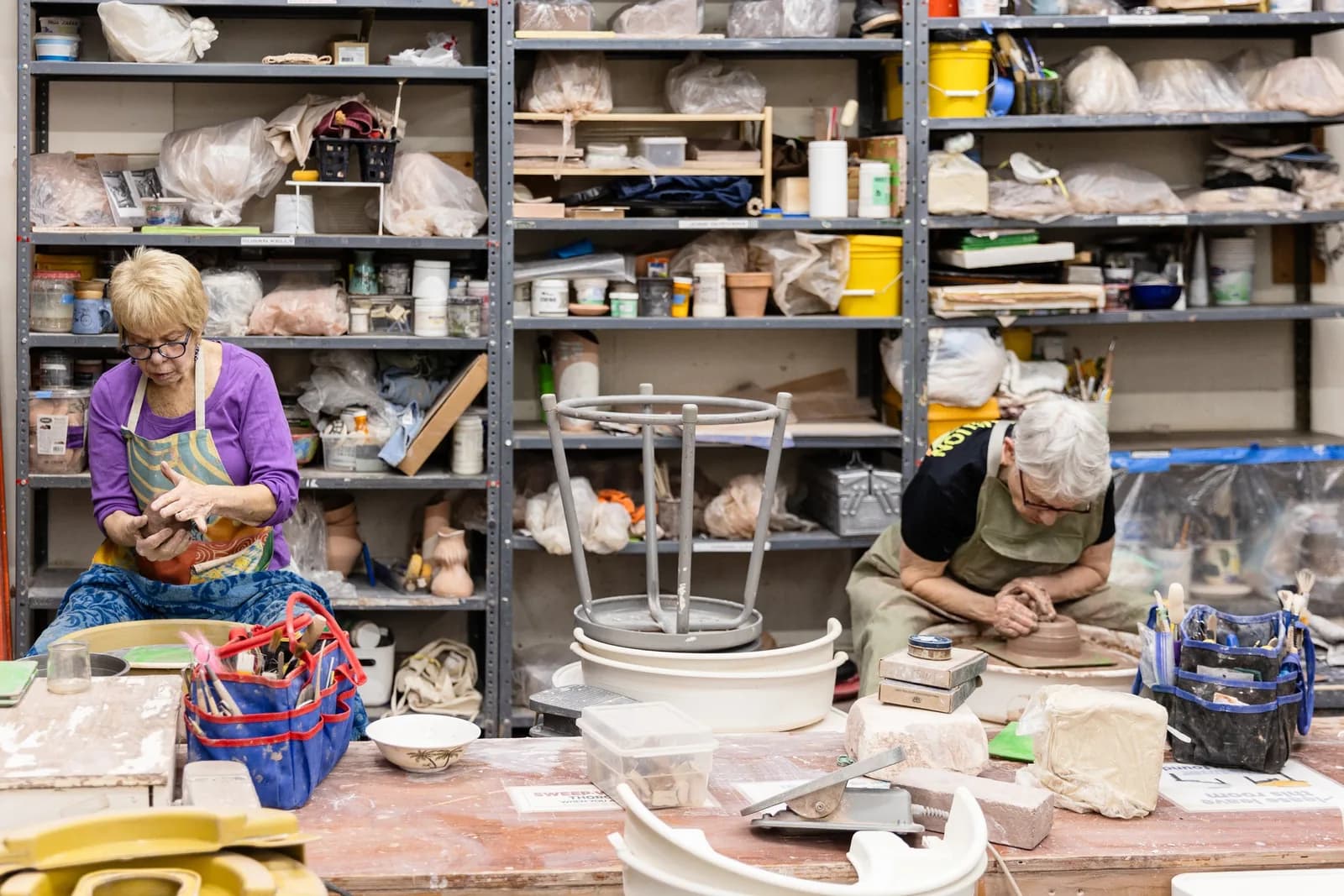 Two potters work at clay stations in a studio with shelves of ceramic supplies and materials