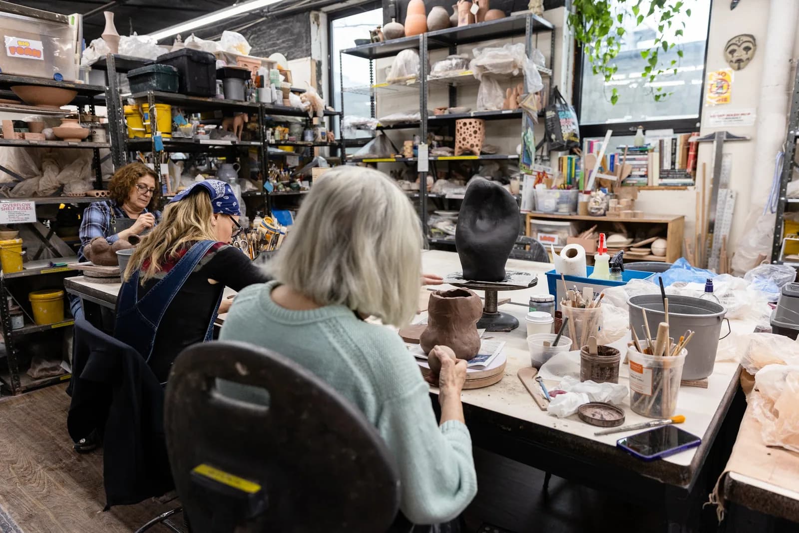 Pottery studio with people working at tables surrounded by clay sculptures, tools, and supplies on shelves