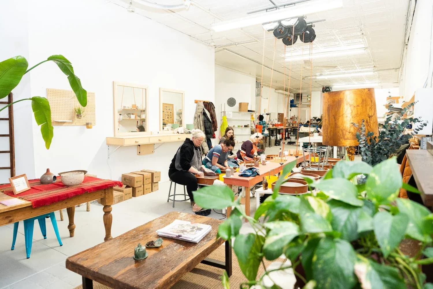 Pottery studio interior with instructors and students working at wooden tables surrounded by clay pieces and plants