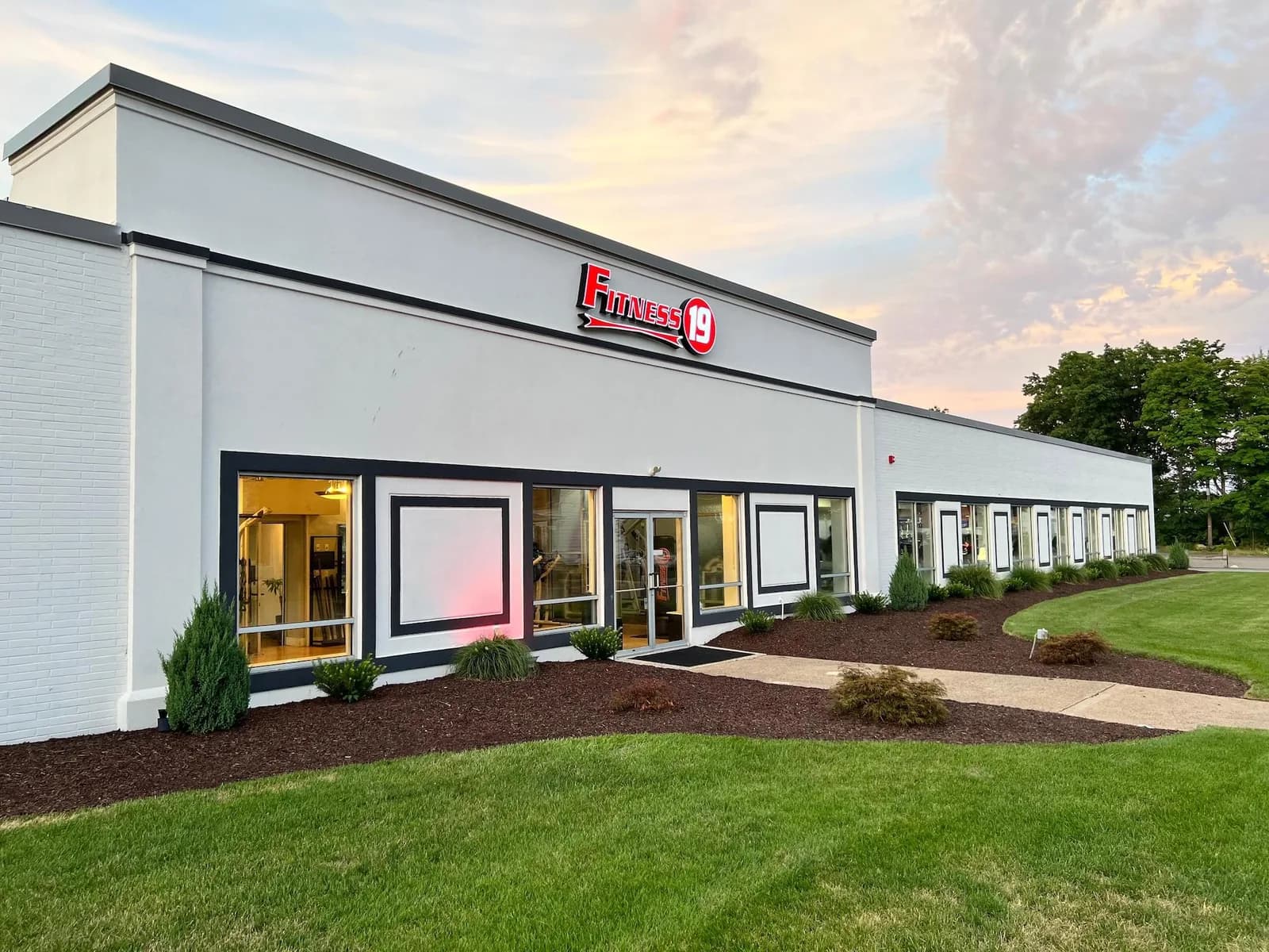Fitness 19 gym exterior with white brick facade, glass entrance doors, and landscaped grounds at dusk