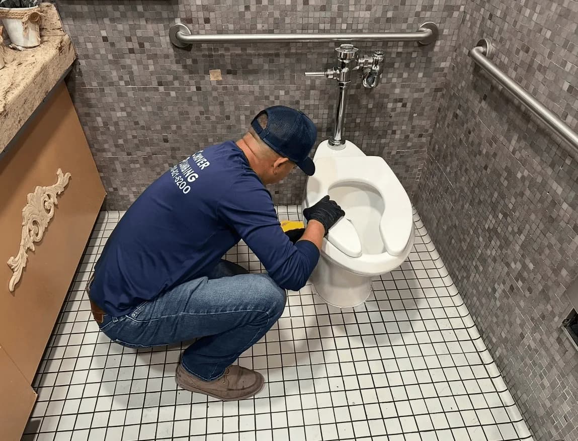 Plumber in navy uniform servicing a white toilet in a gray-tiled bathroom