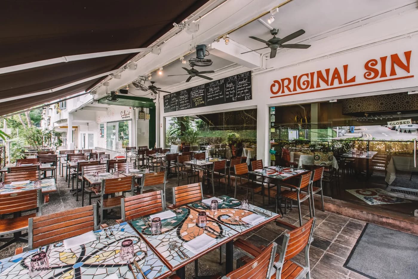 Covered outdoor dining area with orange wooden seating, set tables, and Original Sin storefront