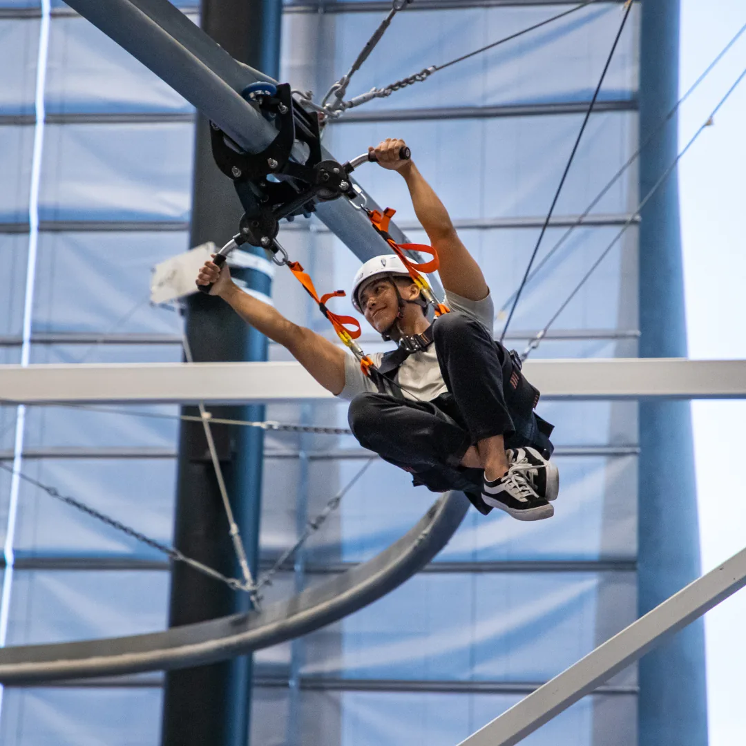 Person zip-lining indoors on a high course with safety gear and helmet