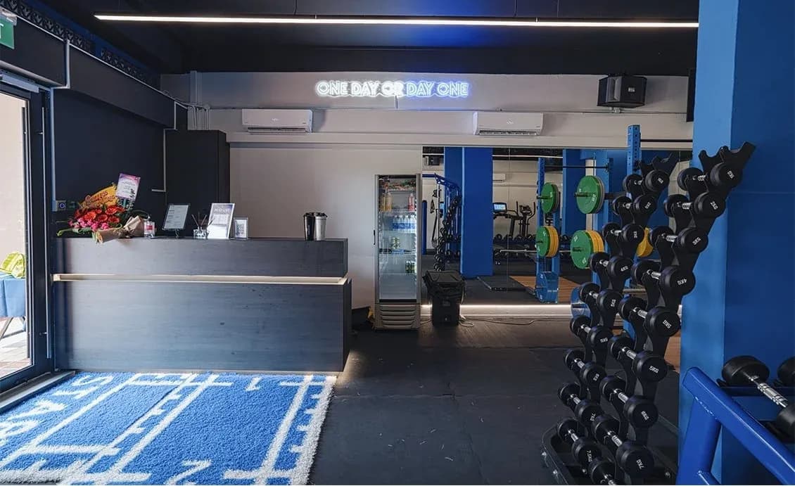 Gym reception area with neon sign, front desk, and organized dumbbell racks with blue-painted walls