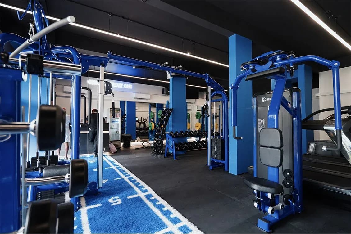 Well-lit gym interior with blue exercise equipment, columns, and machines along a blue-trimmed floor