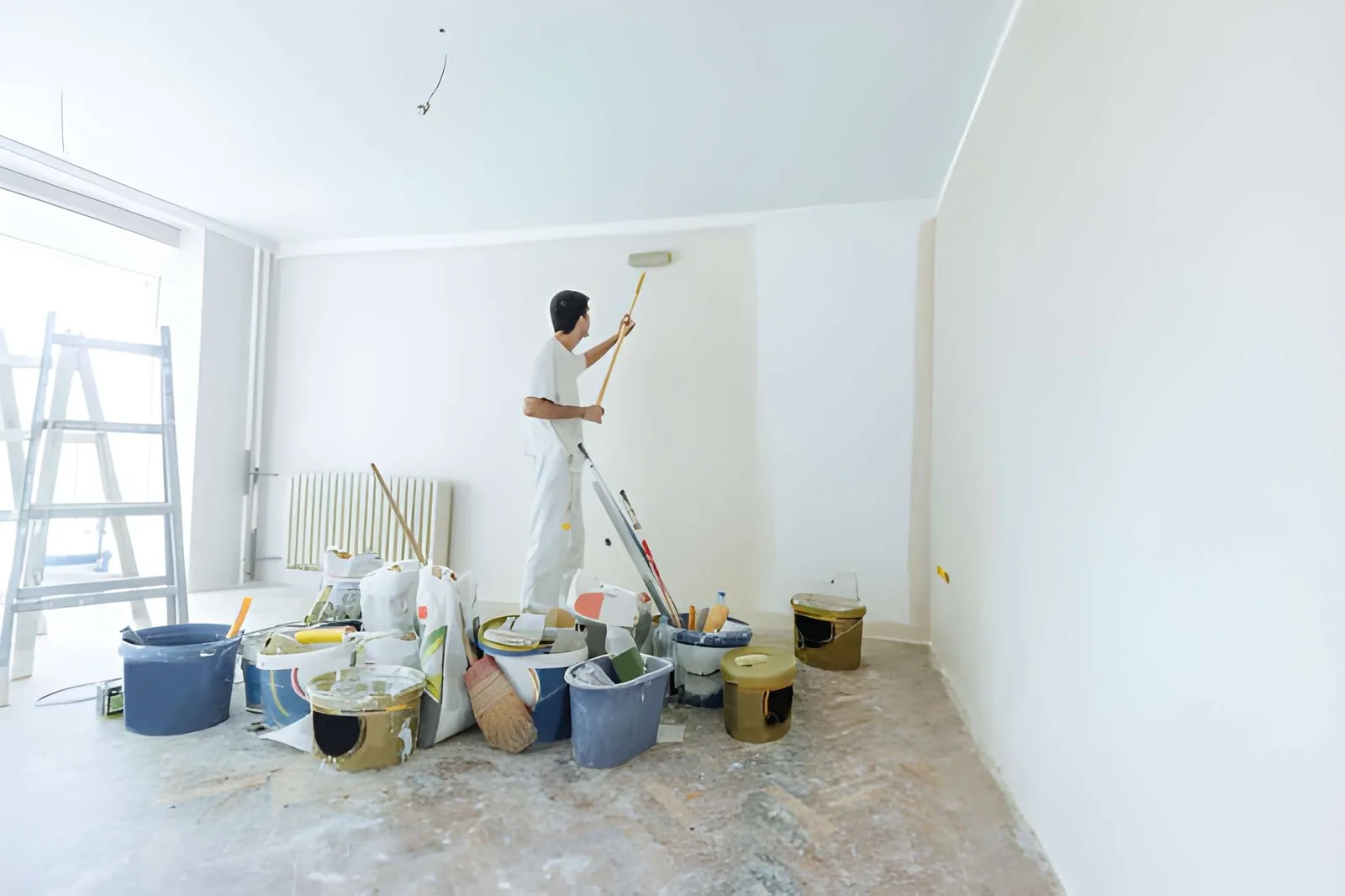 Worker on ladder applying paint or coating to ceiling with paint supplies on floor