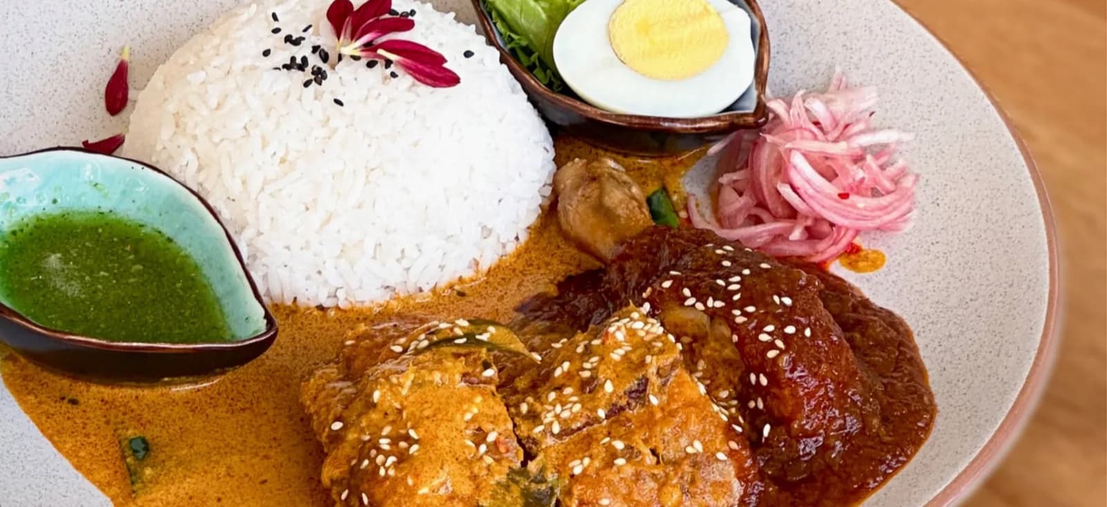 Curry dish with white rice, fried items topped with sesame seeds, pickled red onions, and herb sauce