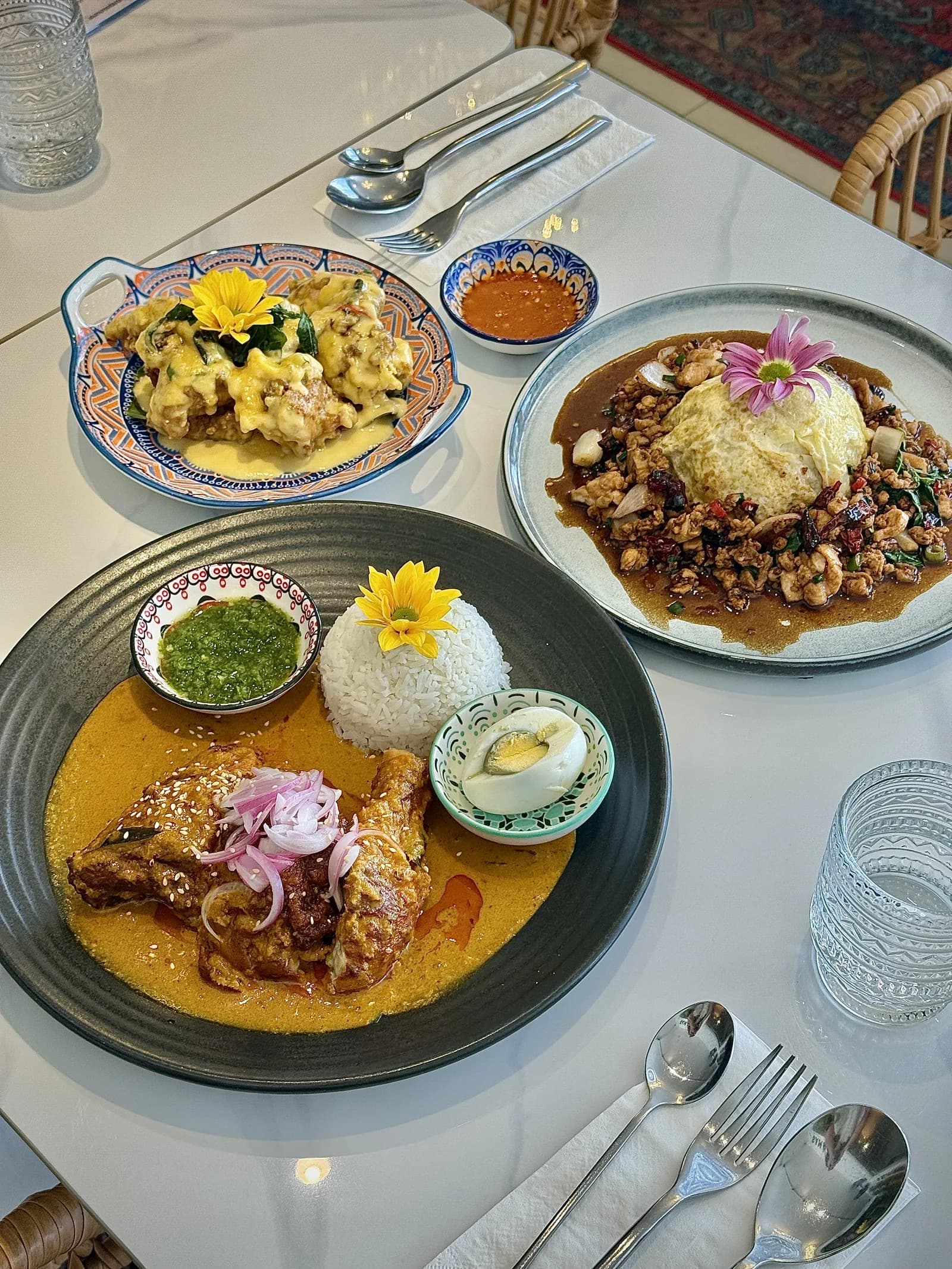 Three plated Malaysian curry dishes with rice, garnishes, and flowers on a white dining table with place settings.
